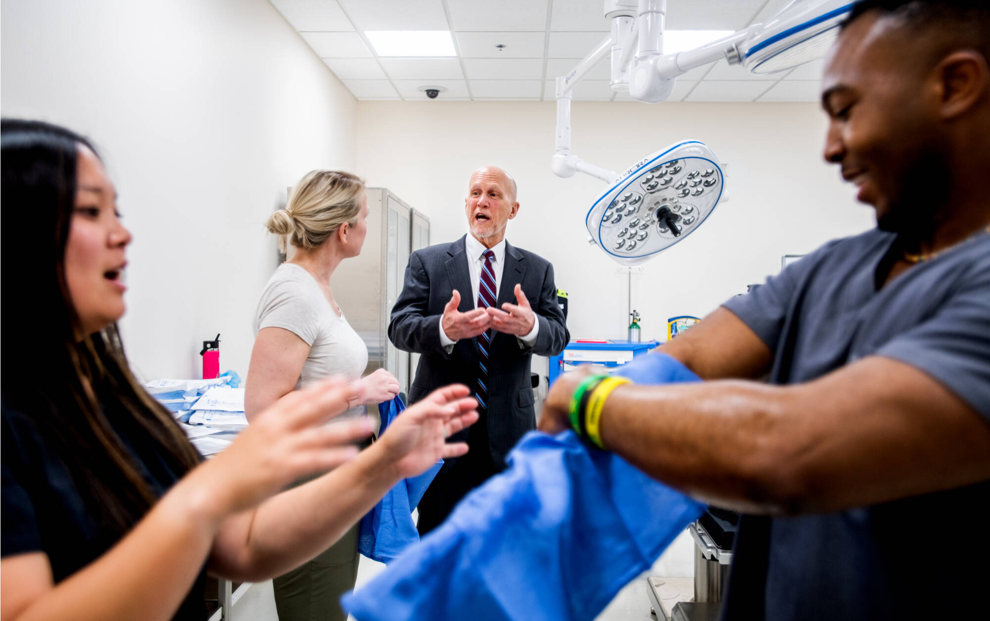 A man in a suit converse with a woman in a medical setting, with surgical lights overhead and a person adjusting scrubs in the foreground.